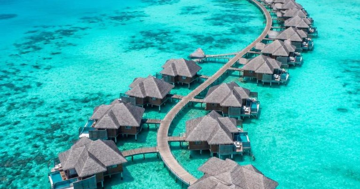A curving line of overwater villas across a turquoise lagoon beside a pale-sand island